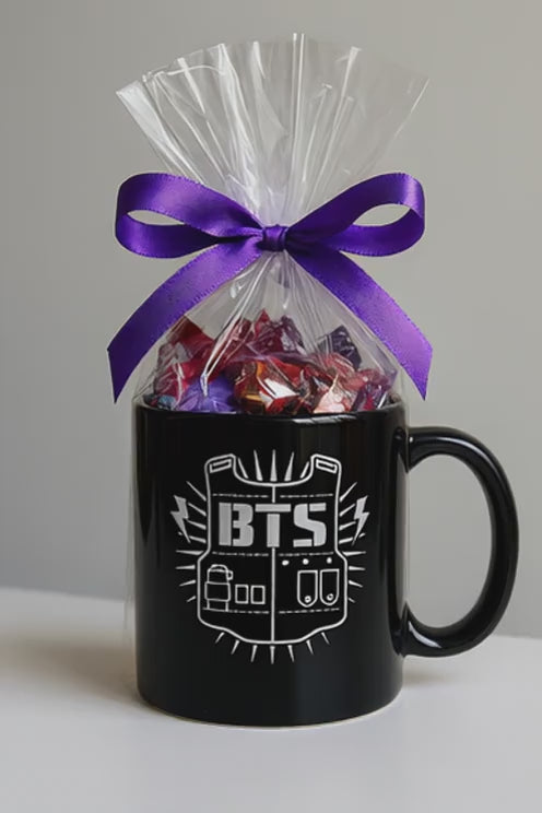 Taza BTS negra con logo bulletproof vest, incluye caramelos surtidos en celofán con lazo púrpura, disponible en SakuPop RD.